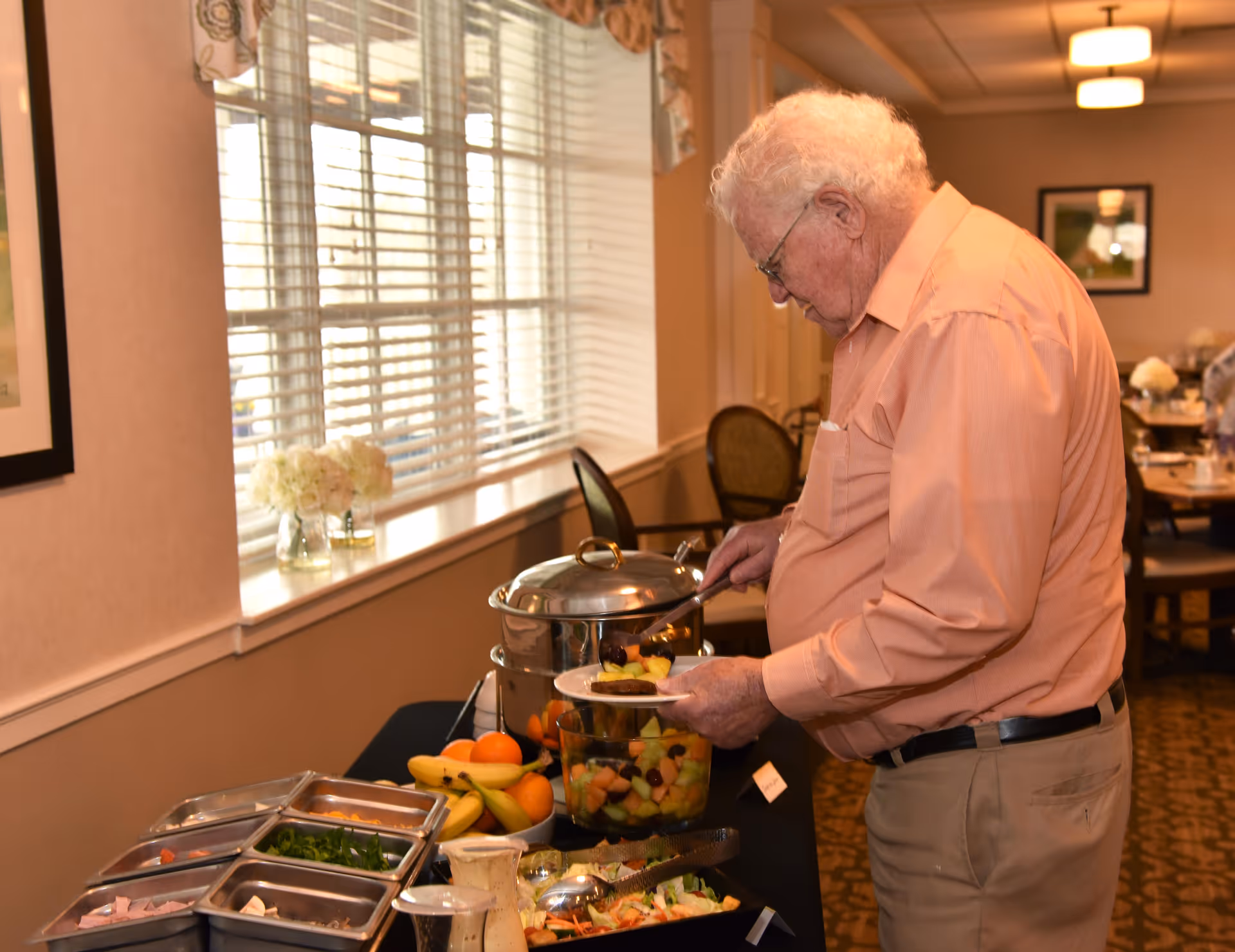An elderly man in a peach shirt and beige pants serving himself fruit salad from a buffet table in a dining room with large windows and floral decorations.