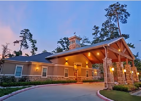 Front entrance of a single-story memory care building with a covered porte-cochere, warm exterior lighting, and surrounding trees at dusk.