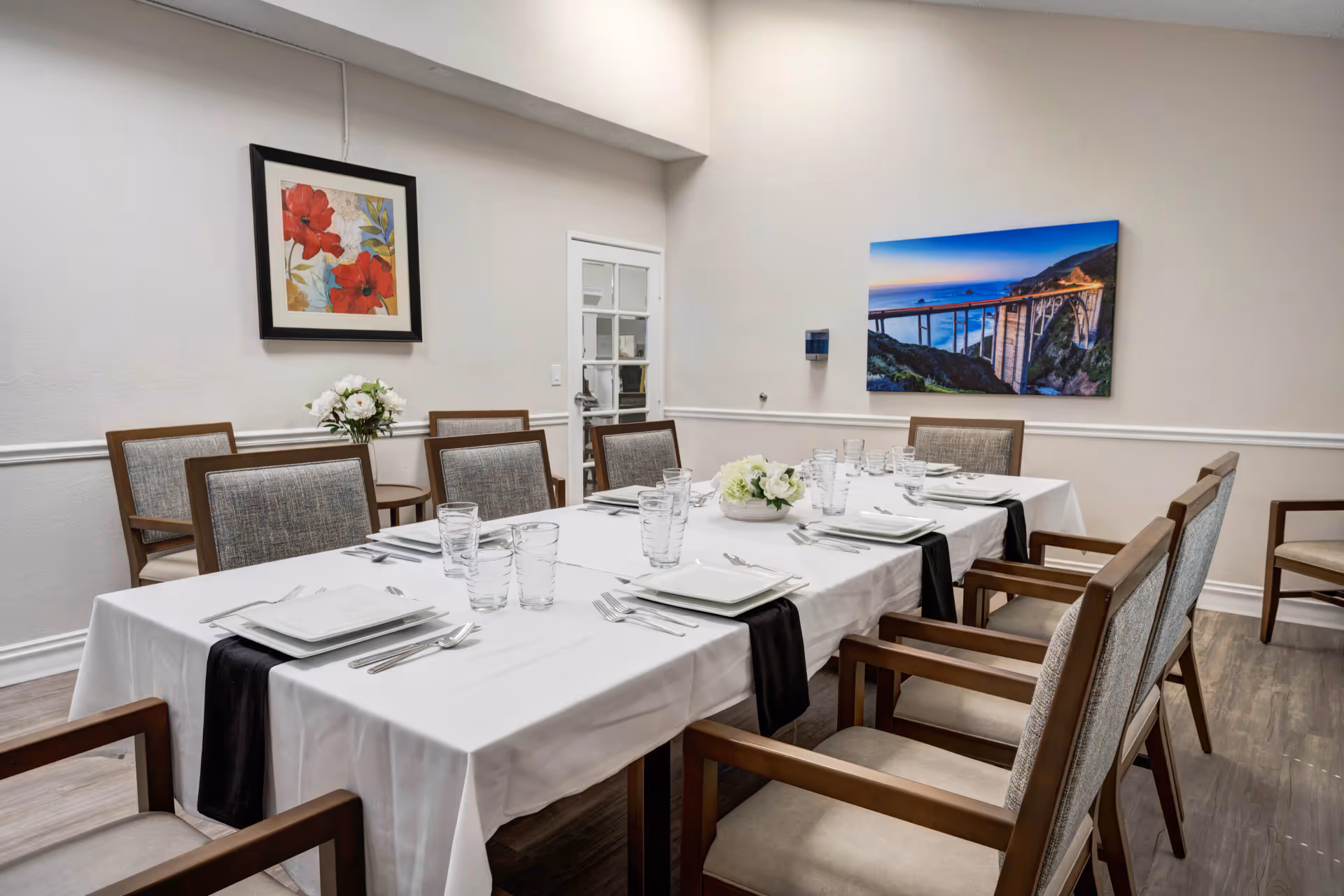 A dining room with a long rectangular table covered with a white tablecloth and black table runners. The table is set with square white plates, silverware, and clear drinking glasses. There are eight wooden chairs with cushioned seats and backs around the table. The room has light-colored walls with two framed pictures hanging, one with red flowers and the other showing a coastal bridge scene. A small round side table with a flower arrangement is visible in the corner.