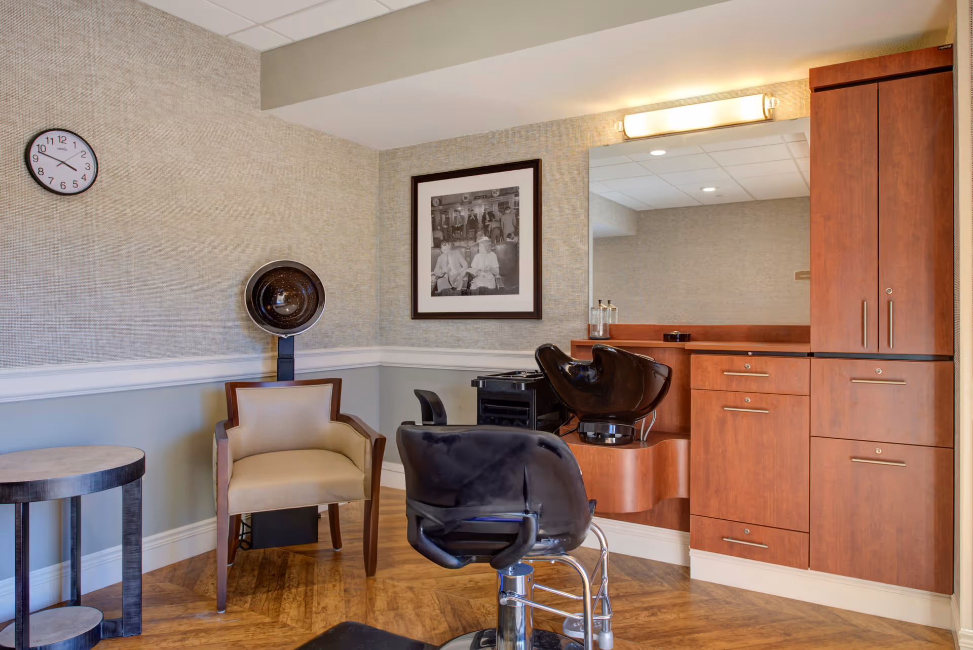 Interior view of a hair salon area in a senior living facility with a black salon chair, a hair washing station, a beige armchair, a round side table, a wall clock, a framed black and white photo, and wooden cabinetry with a large mirror.