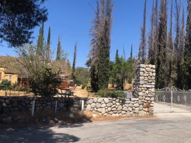 Stone wall and gate entrance to a property with tall trees and a building visible in the background under a clear blue sky.