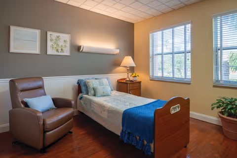 A cozy assisted living bedroom with a single bed covered in a light-colored quilt and a blue throw blanket. Next to the bed is a wooden nightstand with a lamp and a small plant. A comfortable brown recliner chair with a light blue pillow is positioned beside the bed. The room has wooden flooring, two large windows with blinds letting in natural light, and two framed pictures on the wall above the bed.
