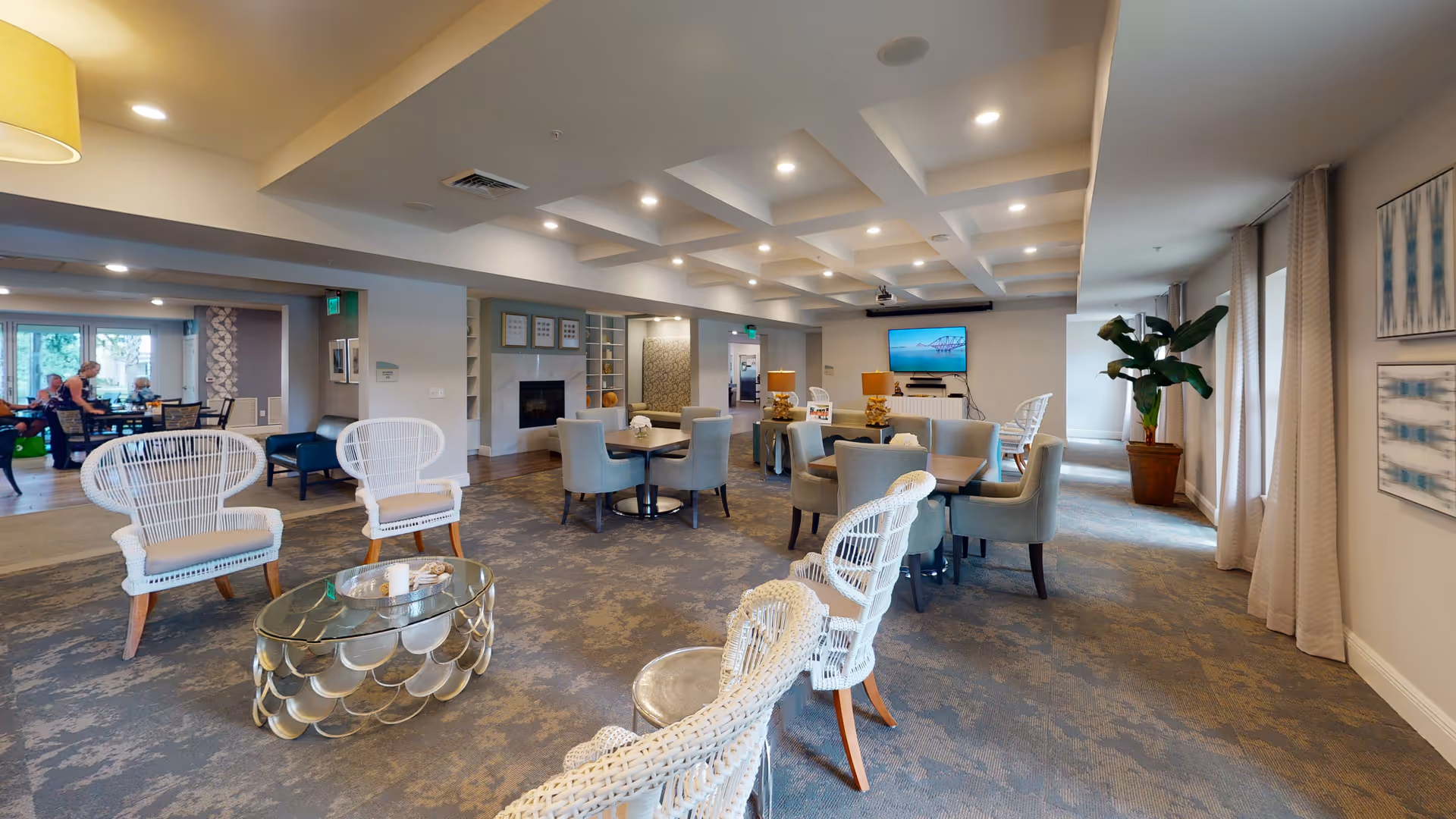 A spacious and well-lit common area in a senior living facility featuring white wicker chairs with cushions arranged around a glass coffee table, several tables with gray upholstered chairs, a fireplace, a wall-mounted TV, and large windows with curtains allowing natural light to enter. The room has a patterned carpet and modern ceiling lighting.