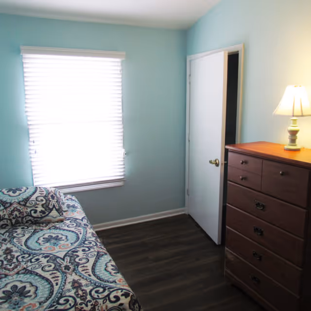 A small bedroom with light blue walls, a window with white blinds, a patterned bedspread on a bed partially visible on the left, and a wooden dresser with a lit lamp on top on the right side. A white door is open, showing a dark room beyond.
