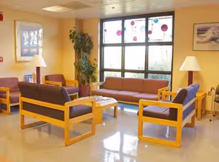A bright senior living lounge with wooden-framed couches, chairs, lamps, and a large window.