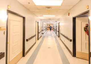 A long, well-lit corridor in a senior living facility with doors lining both sides and a person at the far end.