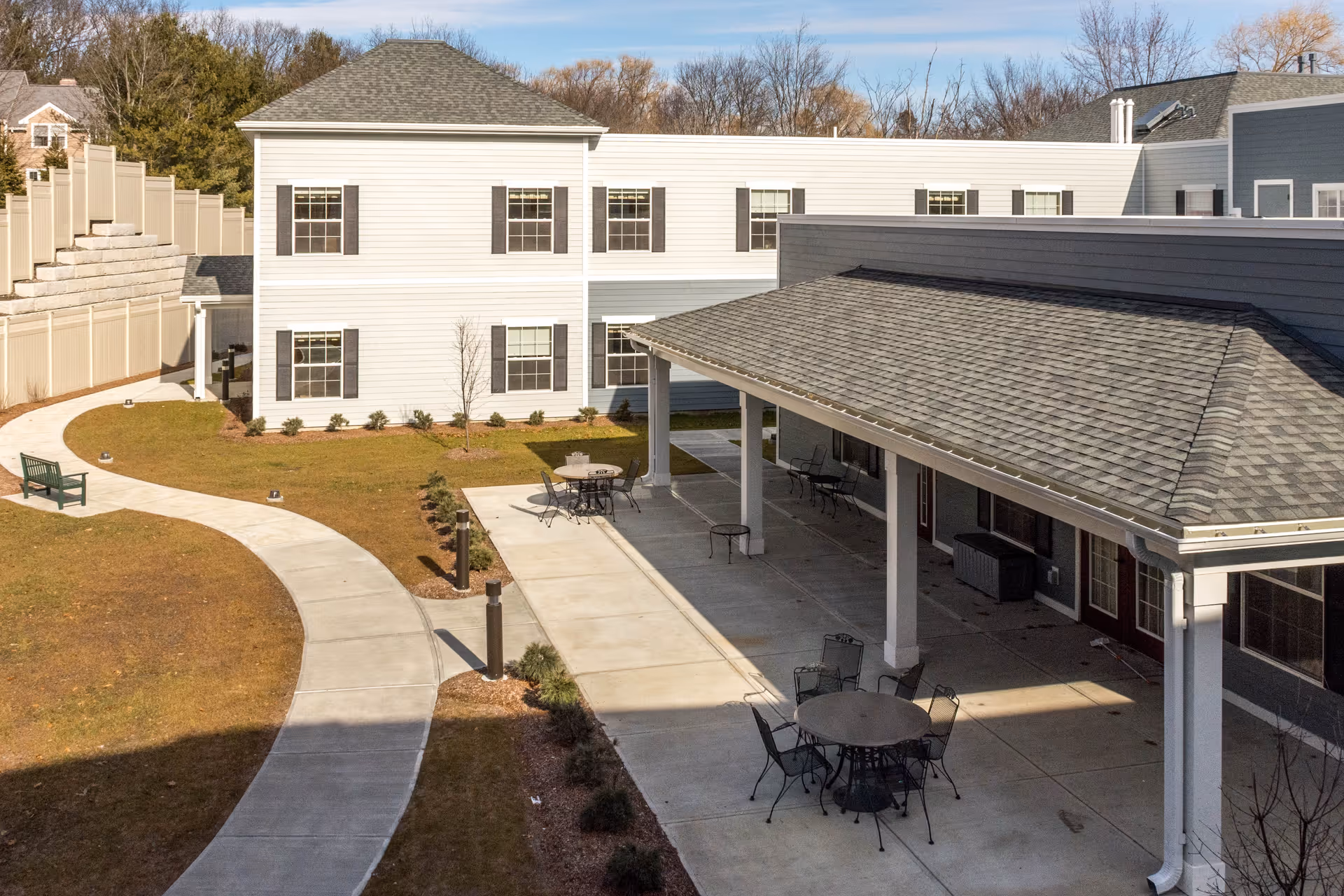 Outdoor patio area at a senior living facility with round tables and chairs under a covered walkway, adjacent to a two-story building with multiple windows and a curved concrete pathway through a grassy lawn.