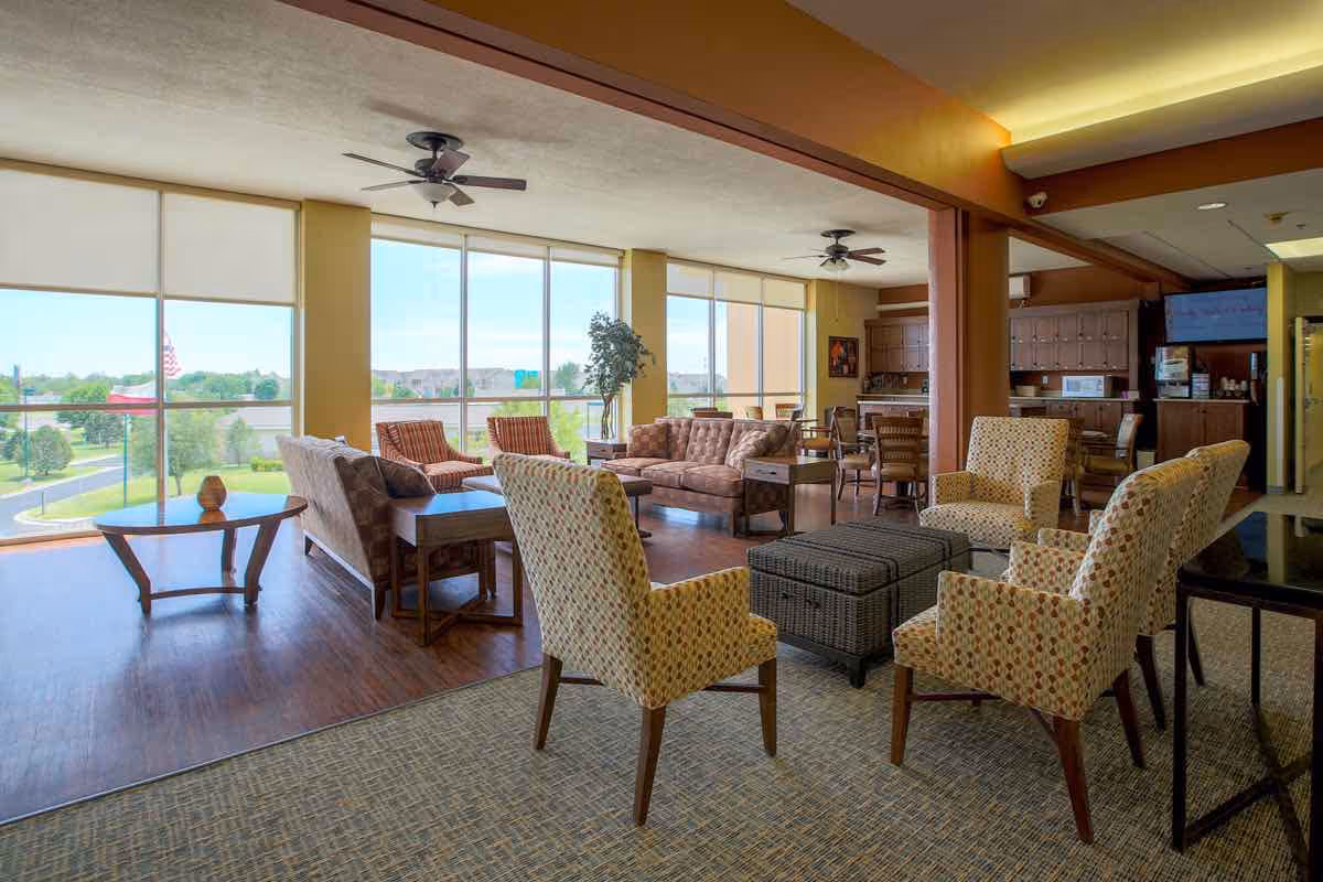 A spacious senior living common area with large windows letting in natural light. The room features multiple seating options including patterned armchairs, cushioned sofas, and wicker chairs arranged around coffee tables. Ceiling fans hang from the ceiling, and there is a kitchenette area with cabinets, a microwave, and a TV in the background. The flooring transitions from carpet to wood near the windows, and a potted plant is placed near the window.
