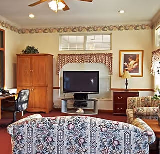 Cozy living room with floral-patterned sofas facing a flat-screen TV, wooden cabinet, desk, and lamp.