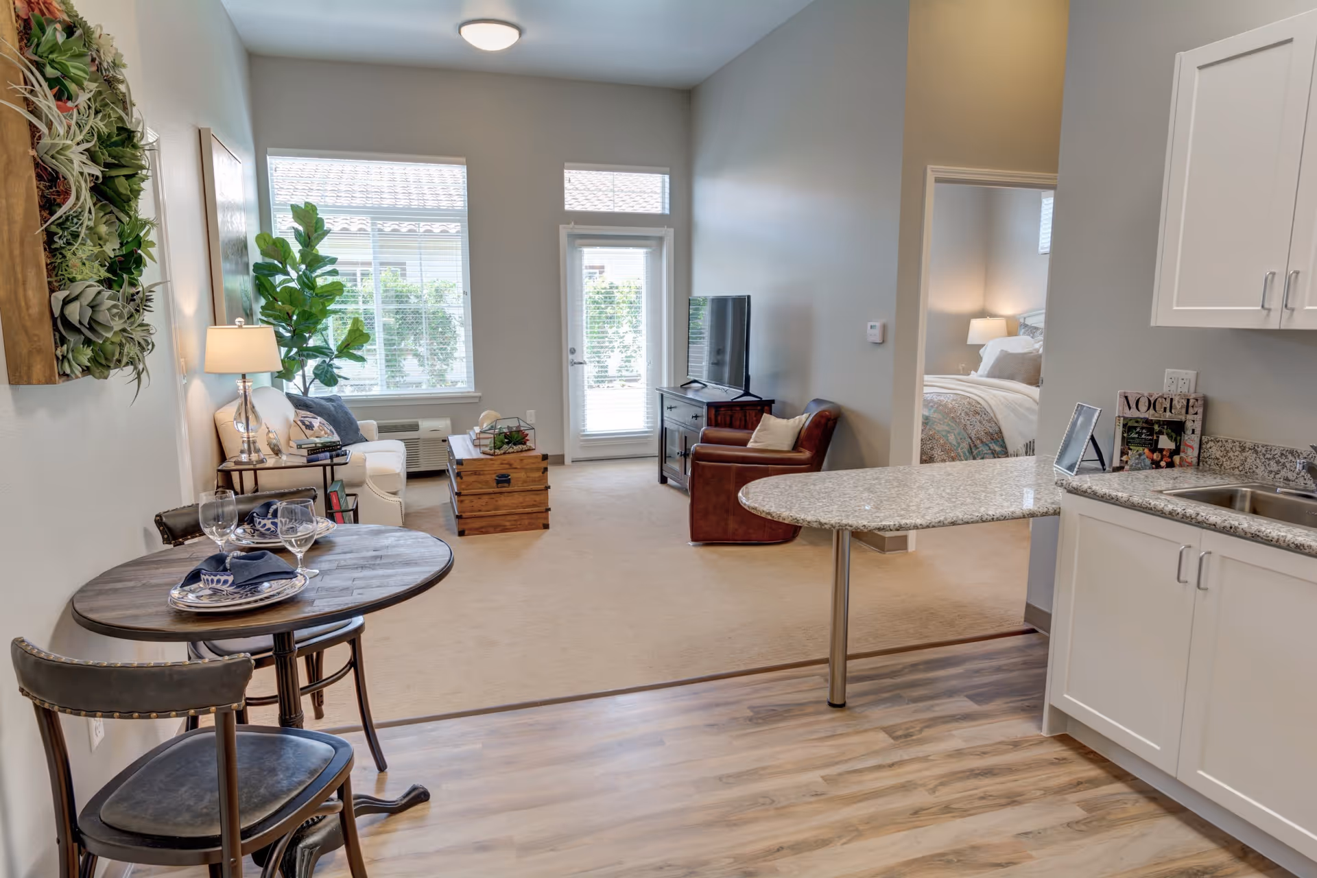 A bright and cozy senior living apartment interior at Rose Arbor Village featuring a small round dining table set for two with plates, glasses, and napkins. The living area has a white sofa, a wooden coffee table, a leather armchair, and a TV on a stand. Large windows and a glass door let in natural light. The kitchen area has white cabinets, a granite countertop with a breakfast bar, and a sink. A bedroom with a made bed and bedside lamp is visible through an open doorway.