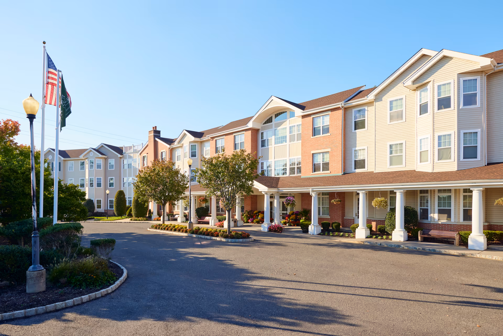 Exterior view of a senior living facility building with a driveway, landscaped garden beds, and three flagpoles displaying the American flag and other flags under a clear blue sky.