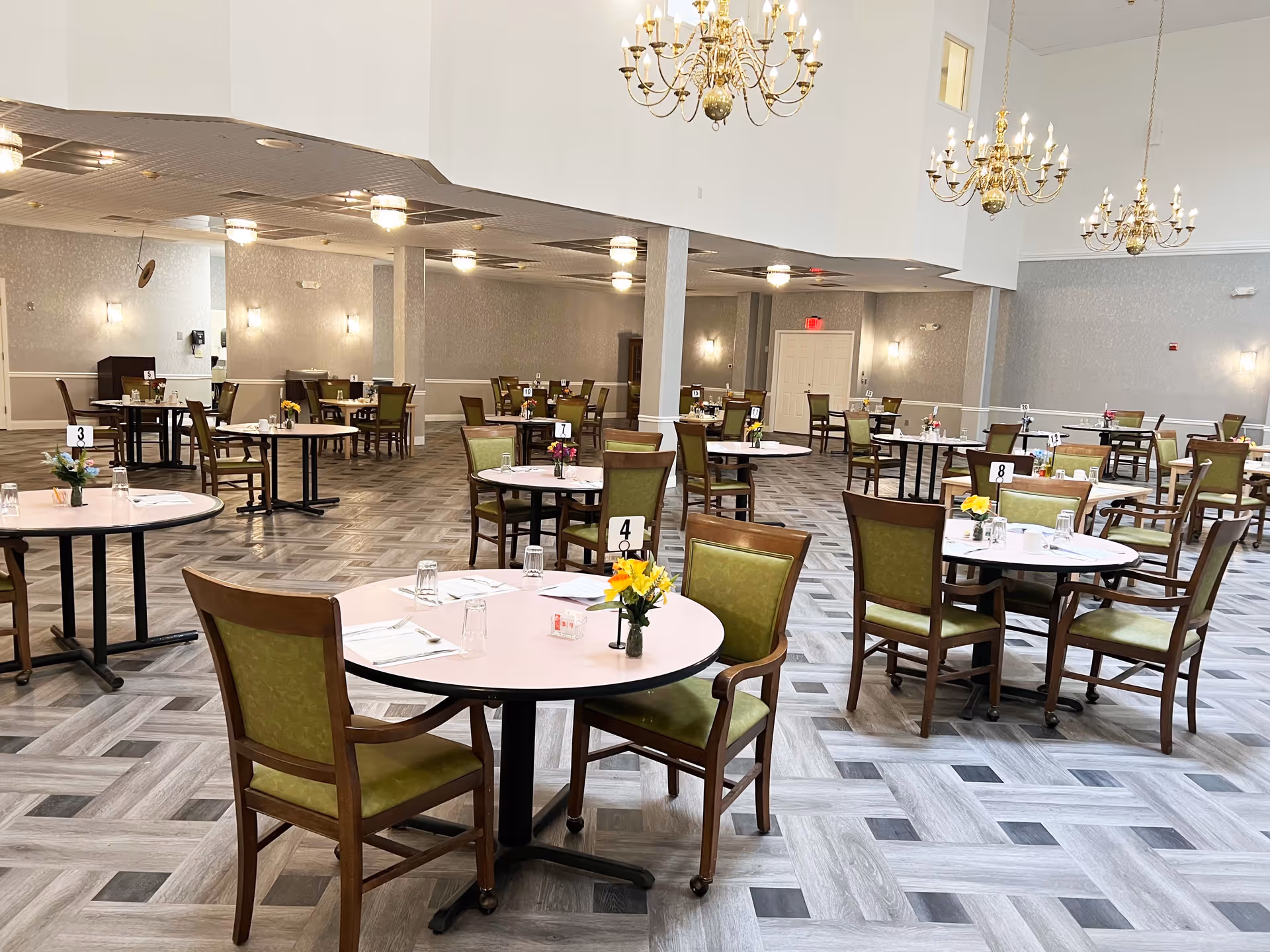 Spacious dining room with multiple round tables, each set with green cushioned chairs, glassware, napkins, and small flower arrangements. The room features a patterned floor, light gray walls, and several ornate chandeliers hanging from a high ceiling.