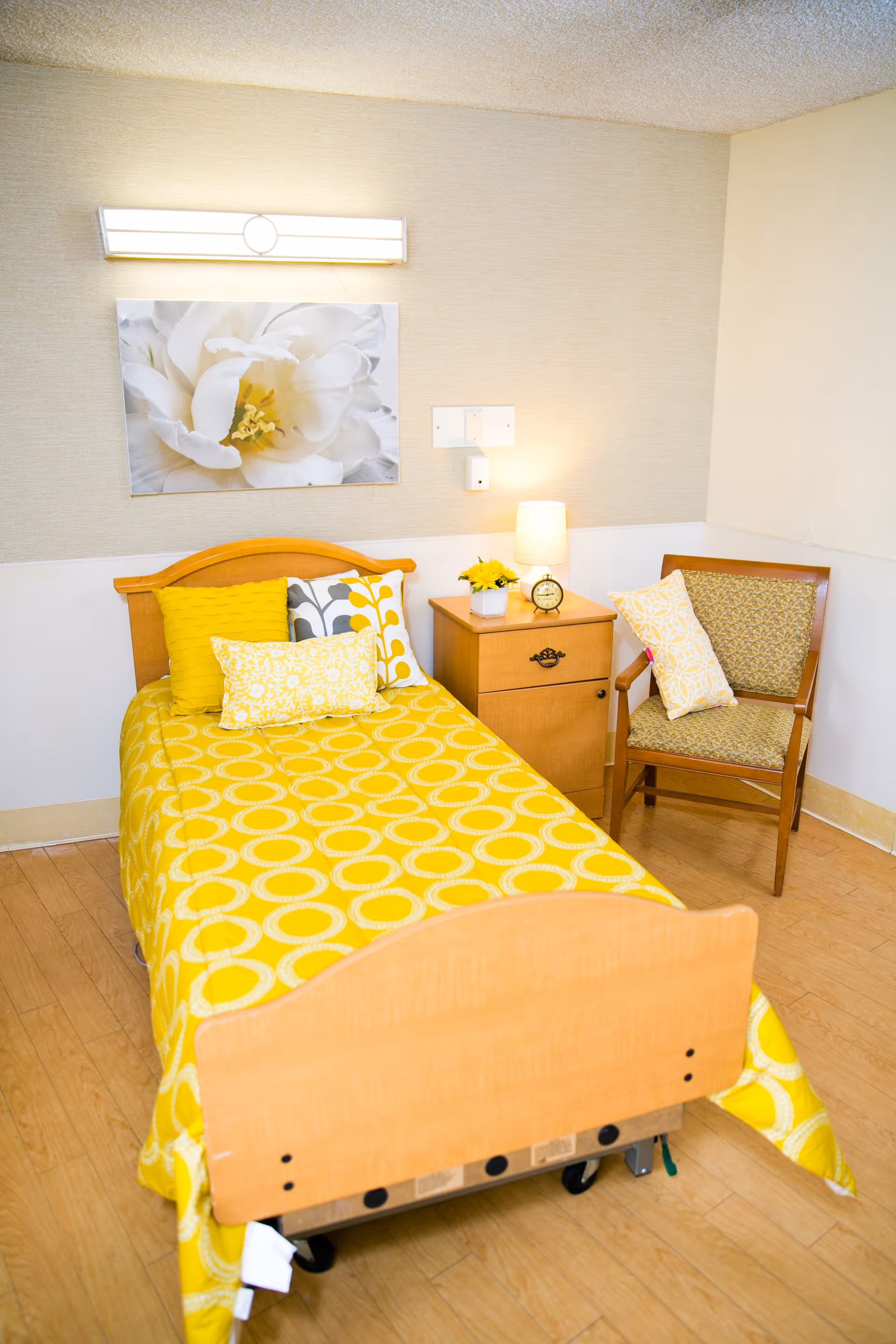 A single bed with a yellow patterned bedspread and multiple decorative pillows in a room. Next to the bed is a wooden nightstand with a small lamp, a clock, and a small flower arrangement. A wooden chair with a cushion and a pillow is placed beside the nightstand. A large white flower artwork is hung on the wall above the bed.