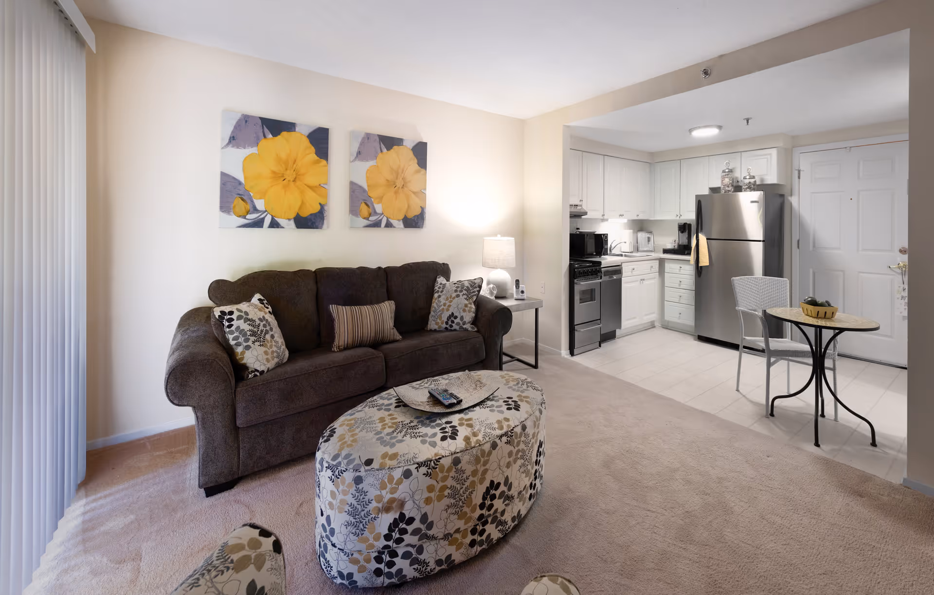 A cozy living area with a brown sofa adorned with patterned cushions, a matching oval ottoman with floral design, and a small side table with a lamp. The room opens into a compact kitchen featuring white cabinets, stainless steel refrigerator, stove, dishwasher, and a small round table with two chairs. Two large yellow flower paintings hang on the wall above the sofa.