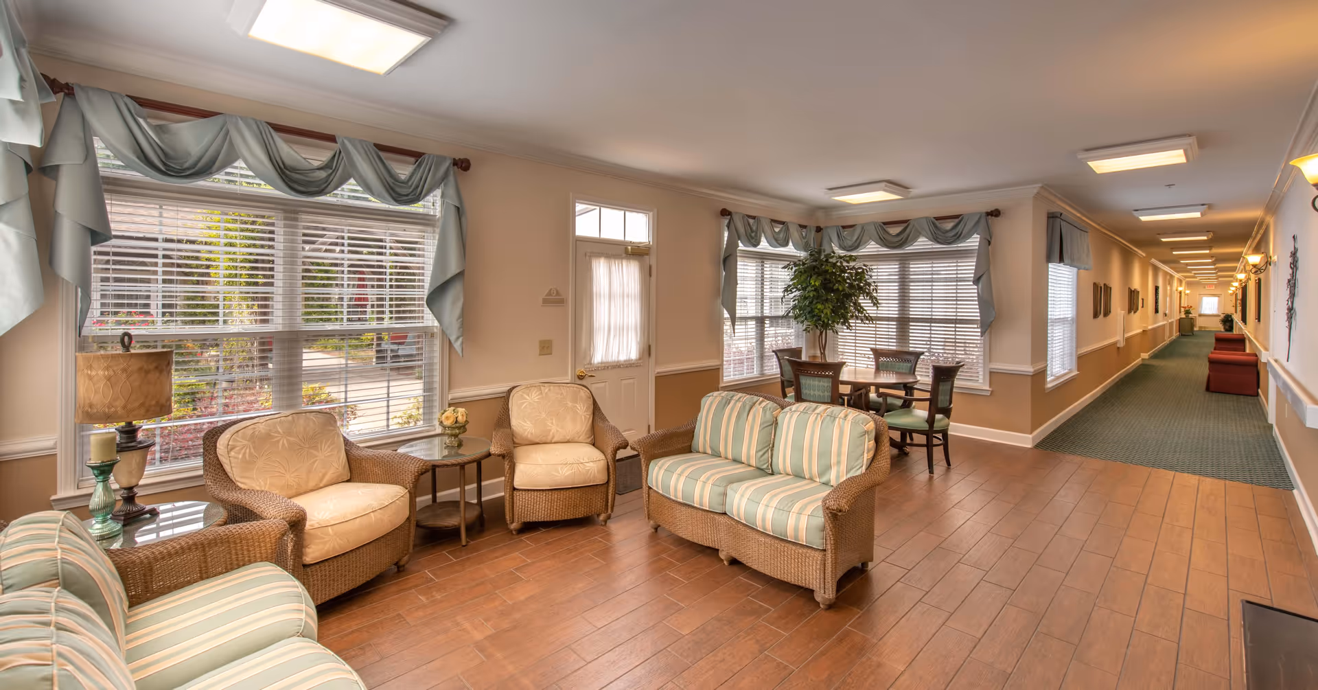 A bright and spacious sitting area in a senior living facility with wicker furniture including a striped loveseat, two armchairs, and a small round table with a lamp and decorative items. Large windows with light blue valances let in natural light. In the background, there is a small dining table with four chairs and a potted plant. A long hallway with carpet and wall decorations extends to the right.