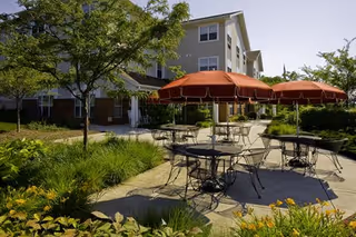 Outdoor patio area with several round metal tables and chairs under large red umbrellas, surrounded by greenery and flowers, with a multi-story senior living building in the background.