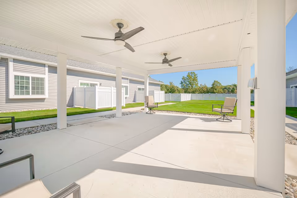 Covered outdoor patio with ceiling fans and seating overlooking a fenced grassy yard.