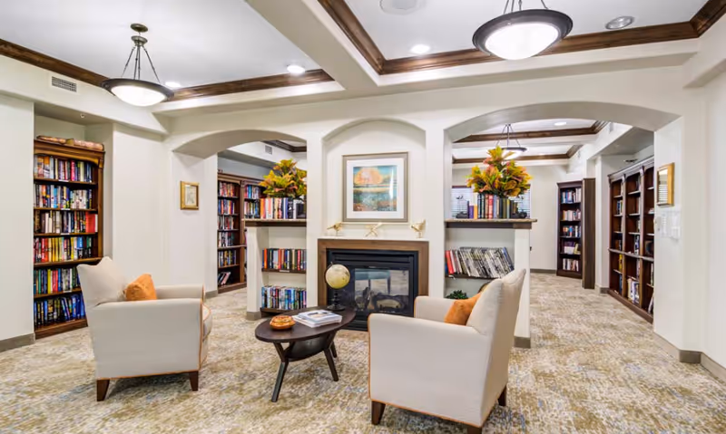 A cozy library or reading room with two beige armchairs facing a small round wooden table. The room features built-in bookshelves filled with books, a fireplace with decorative items on the mantel, and framed artwork above it. The ceiling has wooden beams and recessed lighting, and the carpet has a neutral pattern.