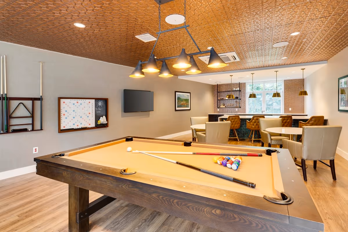 A recreational room featuring a pool table with cues and balls arranged for a game. The room has a decorative ceiling with multiple pendant lights hanging above the pool table. In the background, there is a seating area with round tables and chairs, a bar counter with high stools, and large windows letting in natural light. A wall-mounted TV and a framed picture are also visible on the walls.