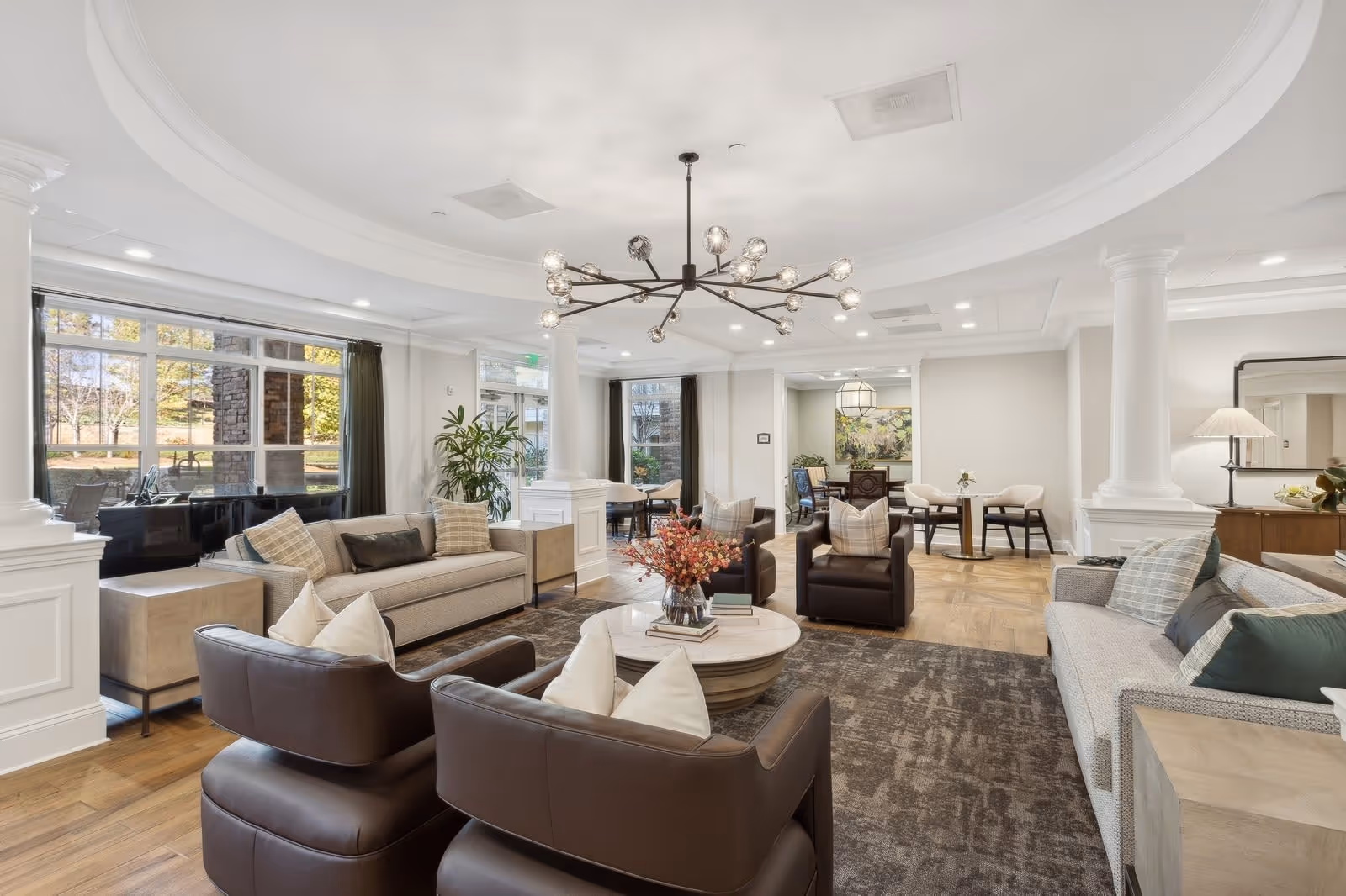 A spacious and well-lit living room area in a senior living facility with multiple seating options including two brown leather armchairs, two beige sofas with cushions, and a round coffee table with a floral centerpiece. The room features large windows with views of outdoor greenery, white columns, a modern chandelier, and a wooden floor with a large area rug. In the background, there are additional seating areas with tables and chairs.