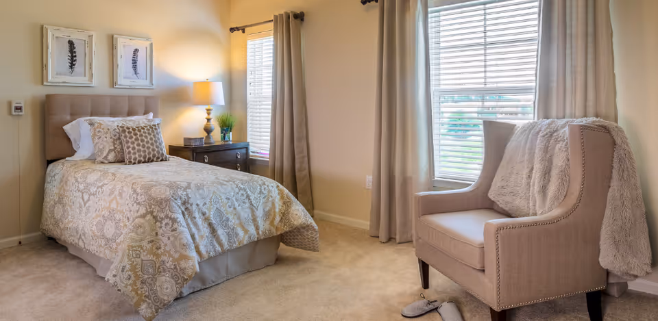 Sunlit furnished bedroom with a single bed, nightstand and lamp, and an armchair by the window with neutral decor.