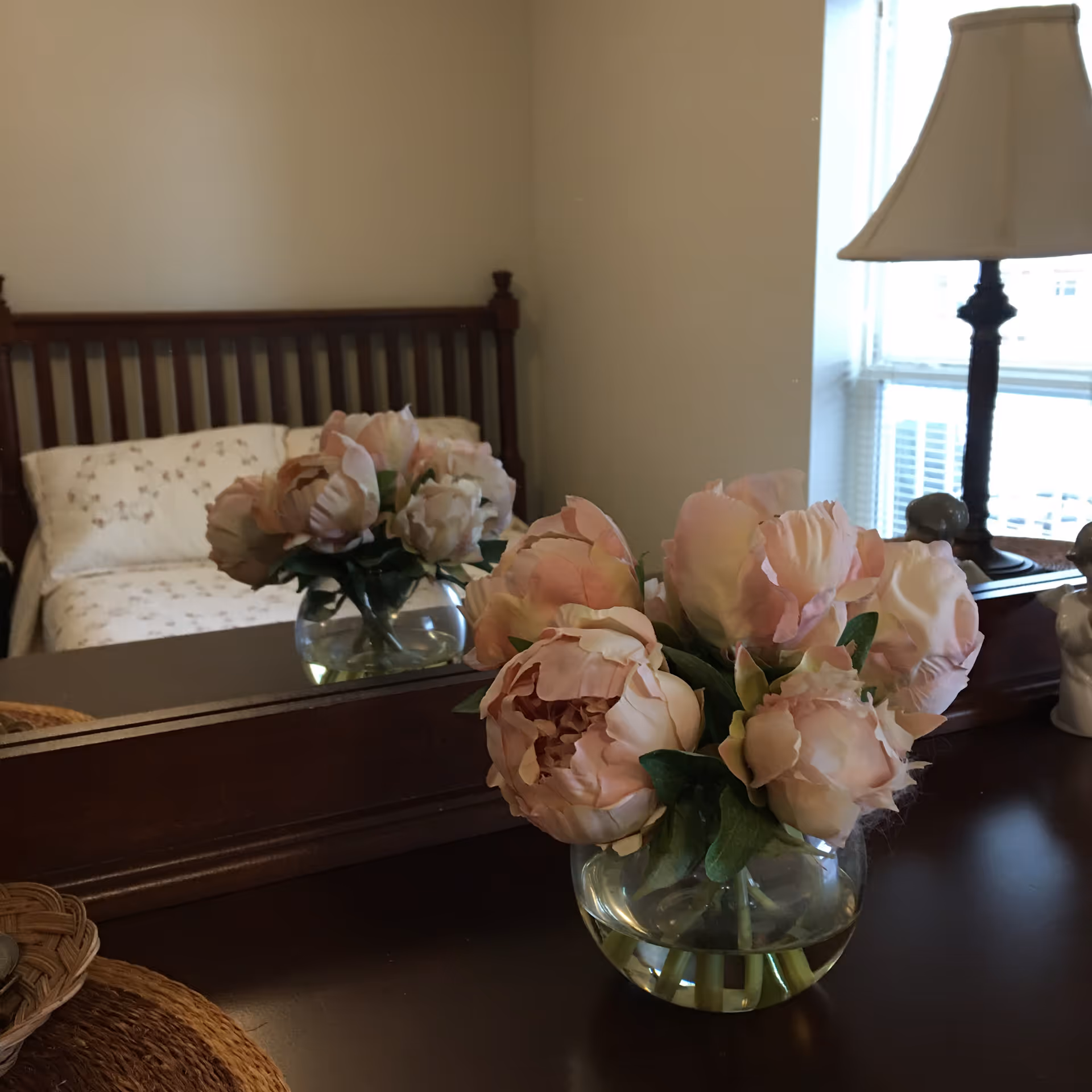 A vase with light pink flowers placed on a dark wooden dresser in front of a mirror. In the reflection, a bed with a wooden headboard and floral bedding is visible, along with a window and a table lamp.