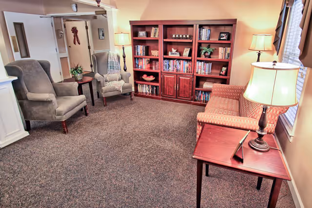 A cozy living room area with two gray armchairs, a wooden bookshelf filled with books and decorative items, a patterned sofa, and three table lamps providing warm lighting. The room has carpeted flooring and a window with curtains on the right side.