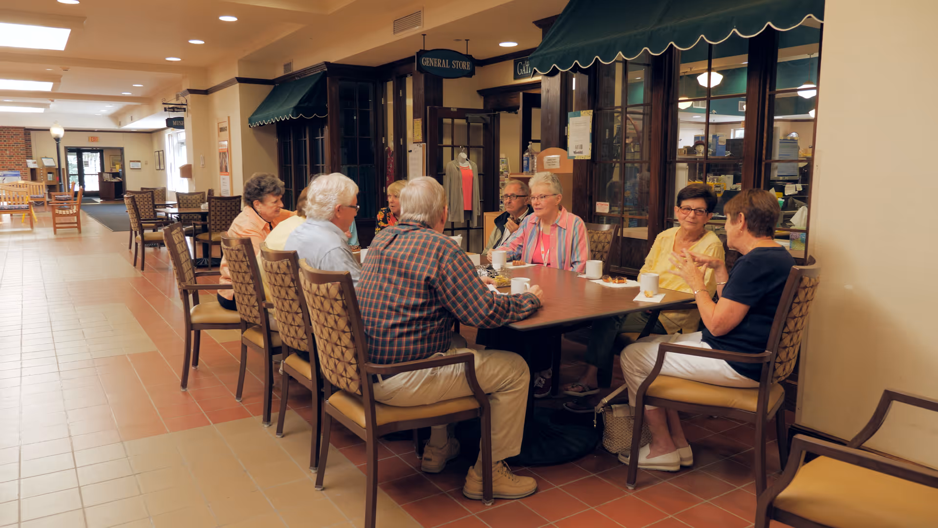 A group of elderly people sitting around a rectangular table in a common area of a senior living facility, engaging in conversation and enjoying drinks. The room has tiled floors, wooden chairs with patterned cushions, and a general store visible in the background.