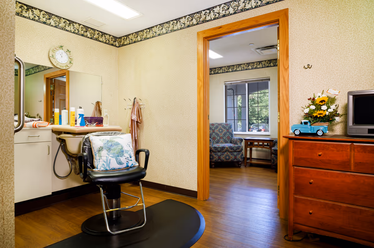 Interior view of a small salon area in an assisted living facility with a black salon chair featuring a turtle-themed pillow, a sink with hair care products on the counter, and a large mirror. The room has beige patterned wallpaper with a floral border near the ceiling. Through an open wooden door, a sitting area with two patterned armchairs, a wooden side table, and a window showing greenery outside is visible. A wooden dresser with a small TV and a decorative blue toy truck with flowers is on the right side.