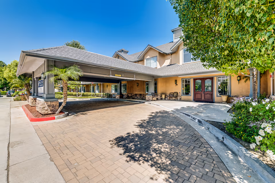 Exterior view of the entrance to Chino Hills Senior Living facility showing a covered drop-off area with stone pillars, a paved driveway, landscaped greenery including palm trees and bushes, and a building with beige walls and a gray roof under a clear blue sky.