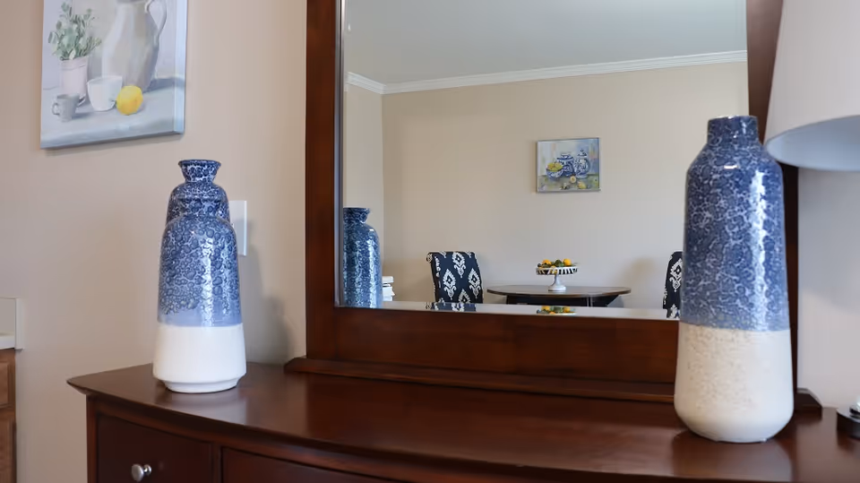 A wooden dresser with a large mirror reflecting a dining area with a round table, two patterned chairs, and a bowl of fruit. On the dresser are two blue and white ceramic vases and a lamp. The walls are beige with framed artwork.