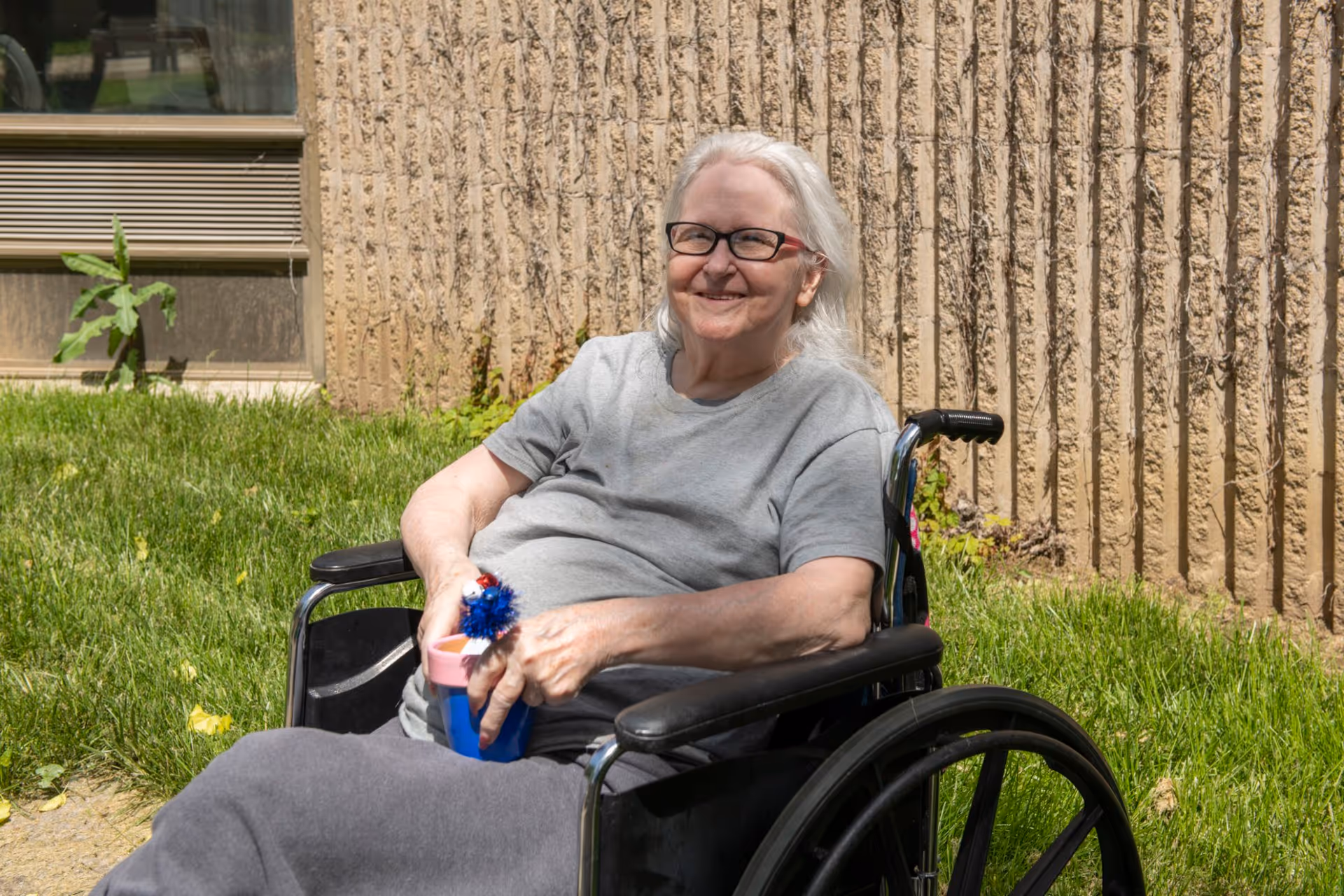 An elderly woman with white hair and glasses sitting in a wheelchair outdoors on grass near a textured beige wall, smiling and holding a blue cup with a decorative red, white, and blue item.