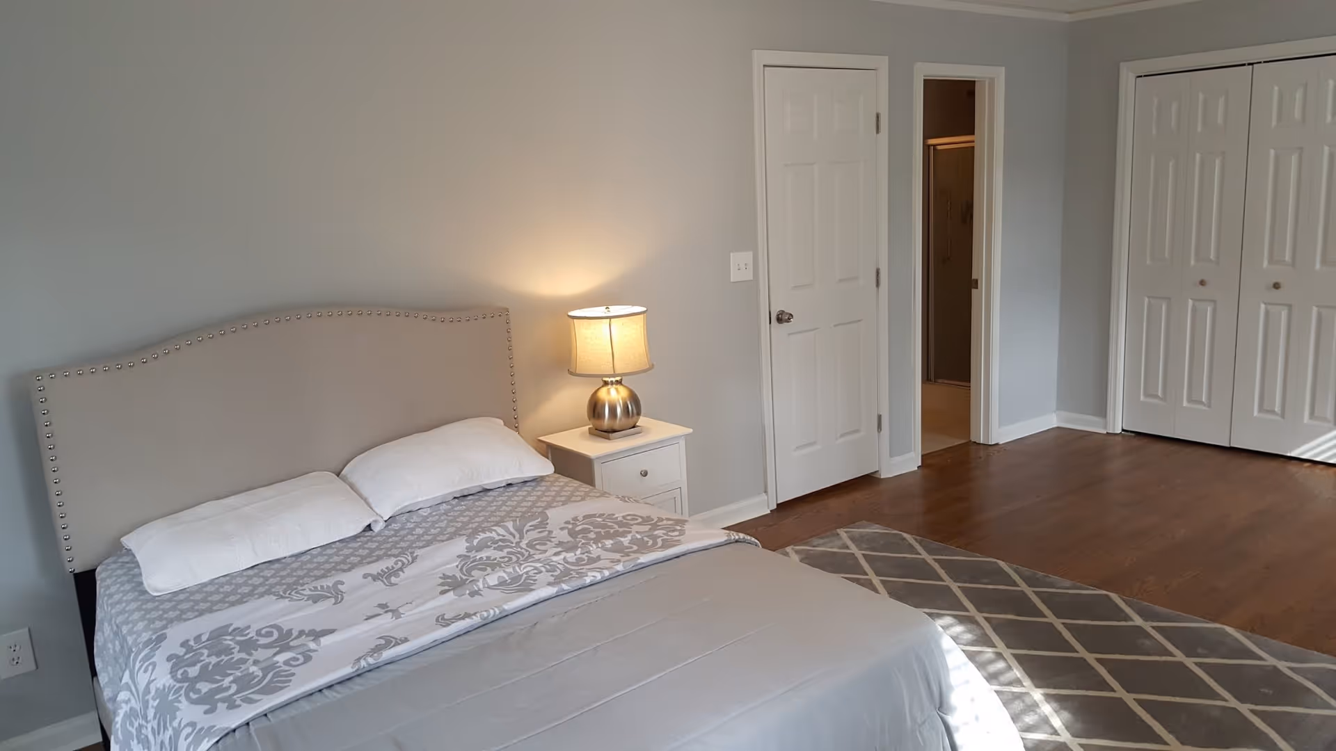 Bright bedroom with a bed, nightstand and lamp, hardwood floors, area rug, and closet and bathroom doors.