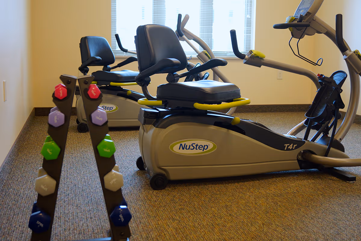 Indoor exercise room with two NuStep recumbent cross trainers and a rack of colorful dumbbells in front of a window with blinds.
