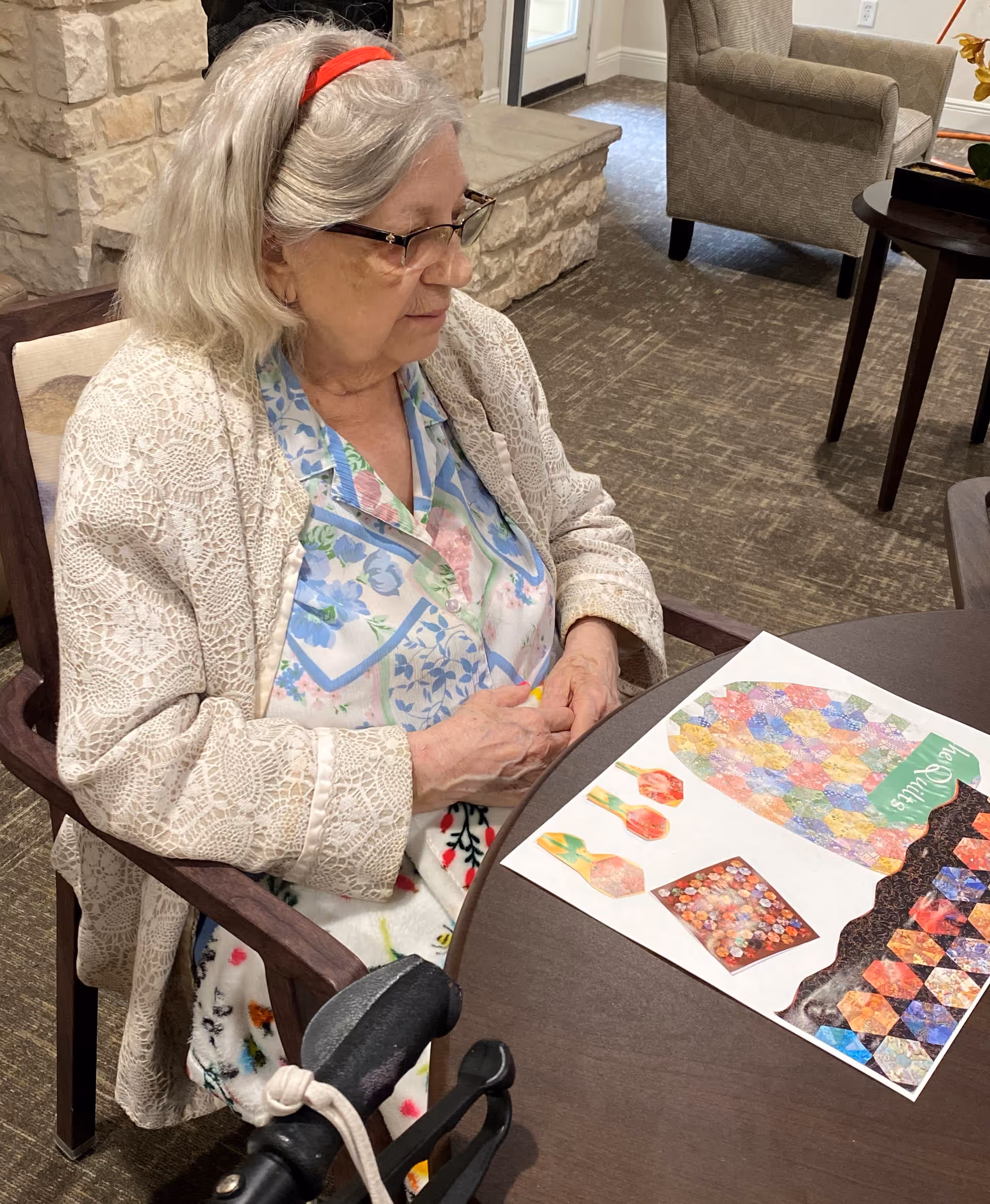 An elderly woman with gray hair and glasses sits at a round table in a cozy room. She is wearing a floral blouse and a cream-colored lace cardigan. On the table in front of her is a colorful paper with various quilt-like patterns. The room has carpeted floors, a stone fireplace, and upholstered chairs.