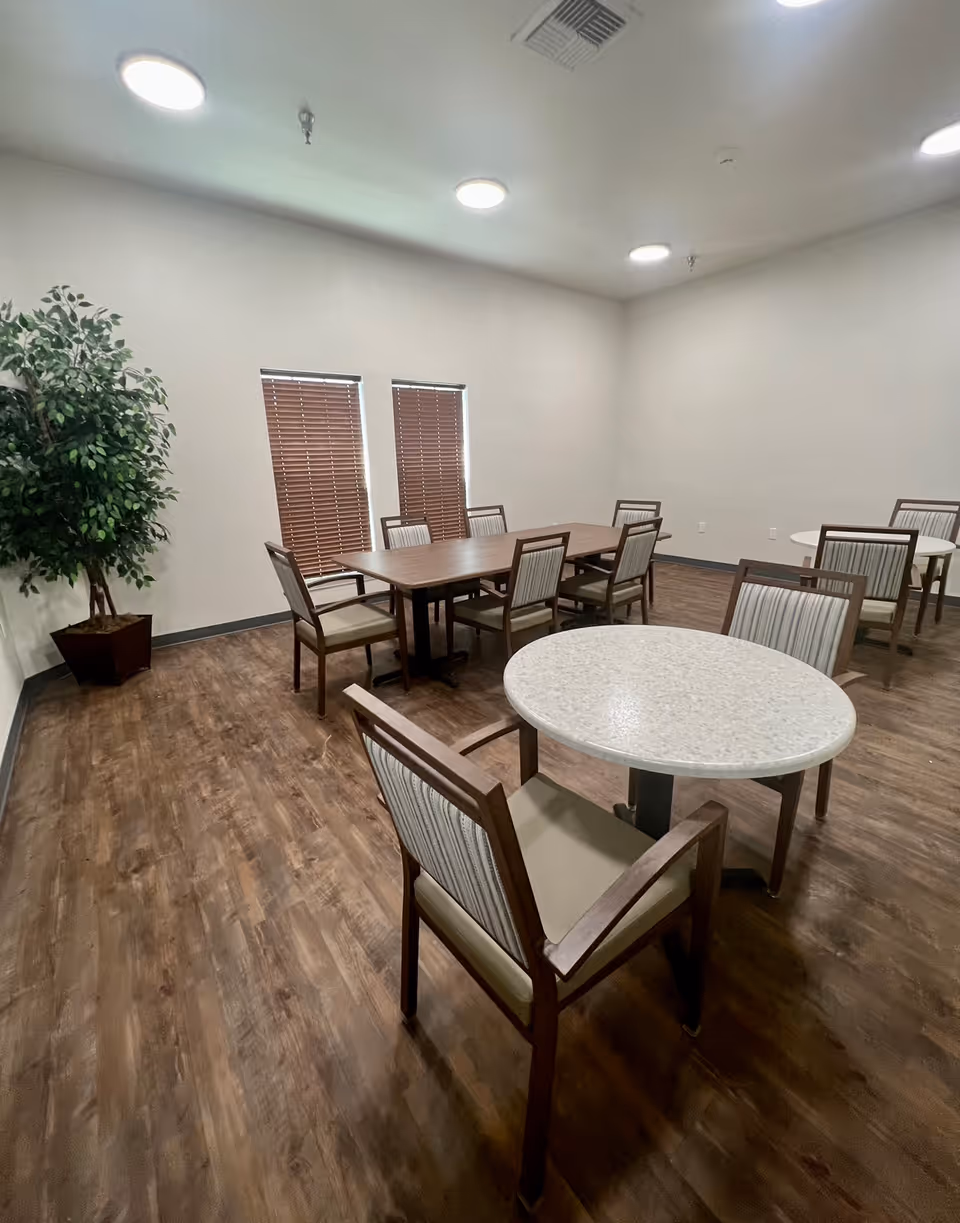 A dining room with wooden flooring, several tables and chairs arranged around the room, two windows with closed blinds, and a large potted plant in the corner. The walls are plain and light-colored with ceiling lights providing illumination.