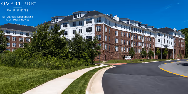 Exterior view of a large multi-story residential building with brick and white siding, surrounded by green grass and trees under a clear blue sky. A curved road and sidewalk lead up to the building. Text on the image reads 'OVERTURE FAIR RIDGE 62+ ACTIVE INDEPENDENT APARTMENT HOMES'.