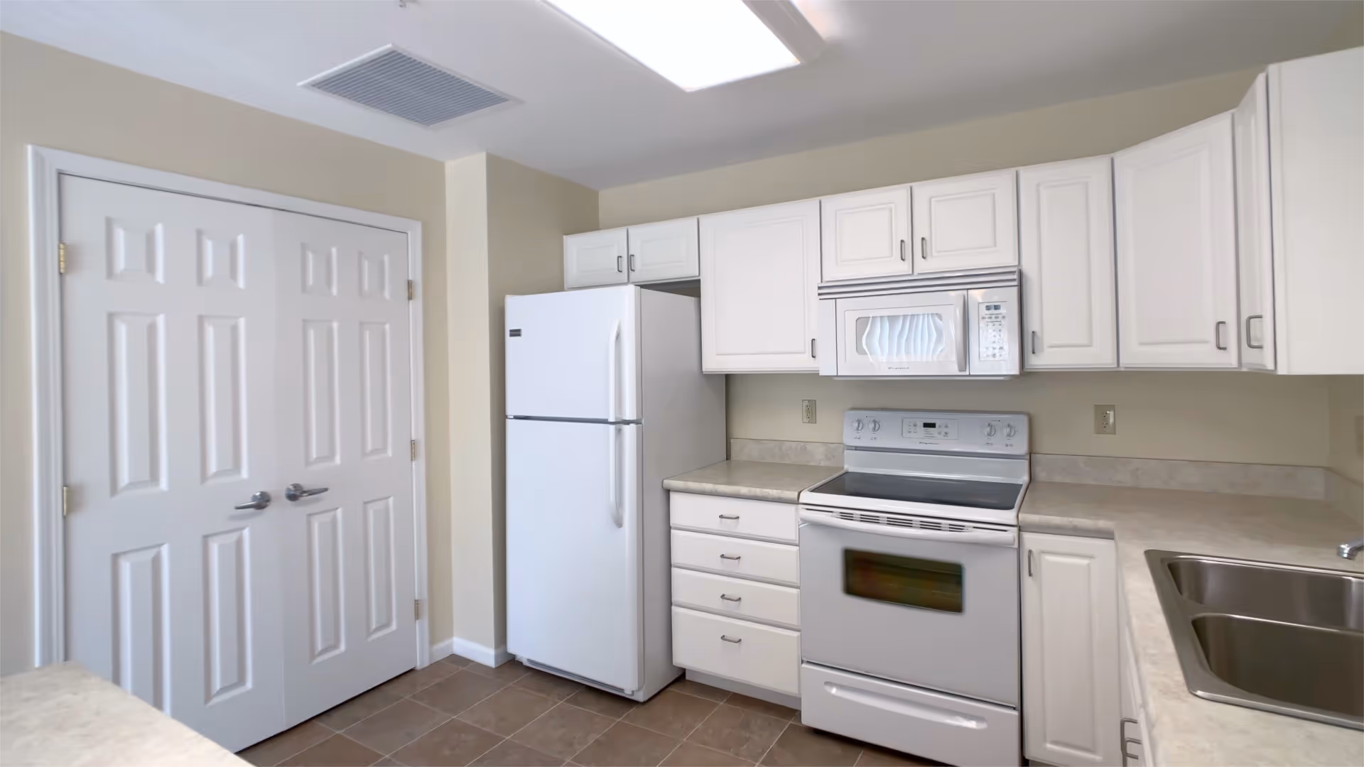 A clean kitchen with white cabinets, a white refrigerator, a white electric stove with an oven, a white microwave above the stove, and a double stainless steel sink. There is a double-door pantry on the left side and beige countertops with a beige tiled floor.