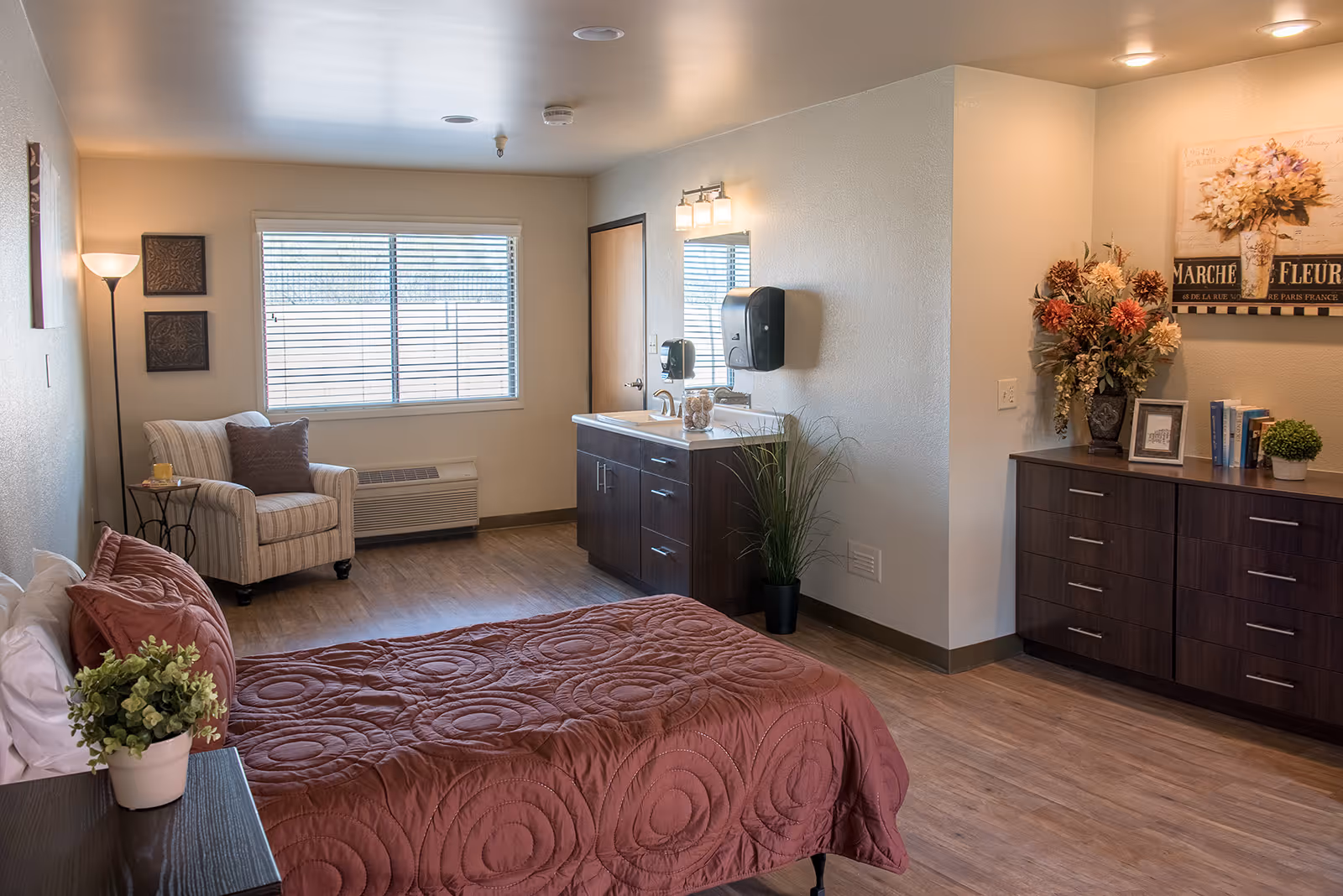 Furnished bedroom with a bed covered by a red quilt, an armchair by the window, dresser cabinets, and a sink/vanity area.