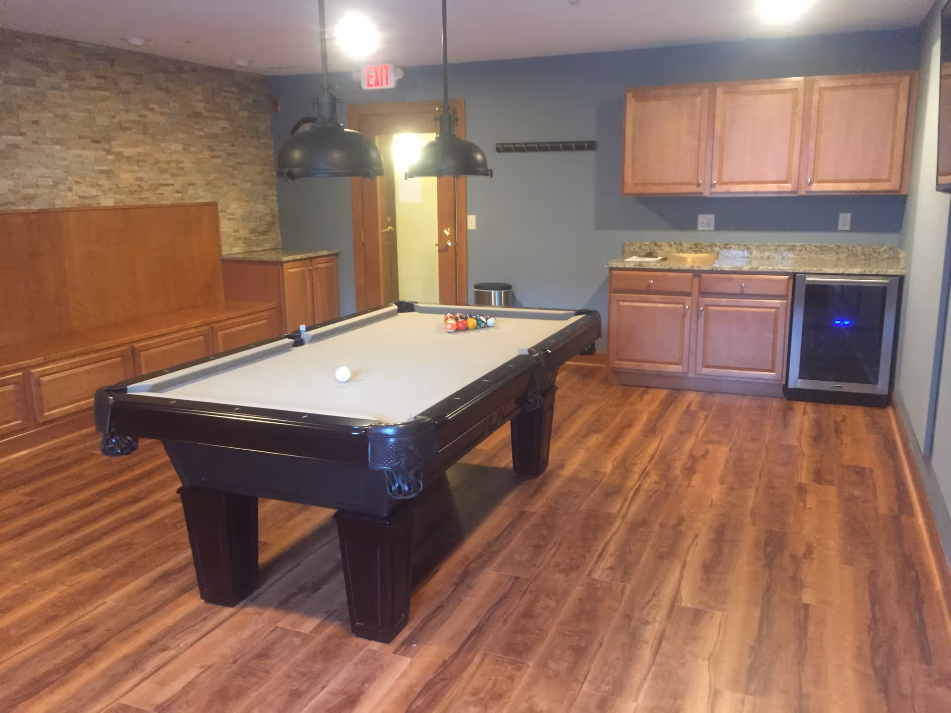 Interior room with a pool table in the center, wooden flooring, a stone accent wall with built-in wooden bench seating, overhead pendant lights, and a kitchenette area with wooden cabinets, granite countertop, and a small refrigerator.