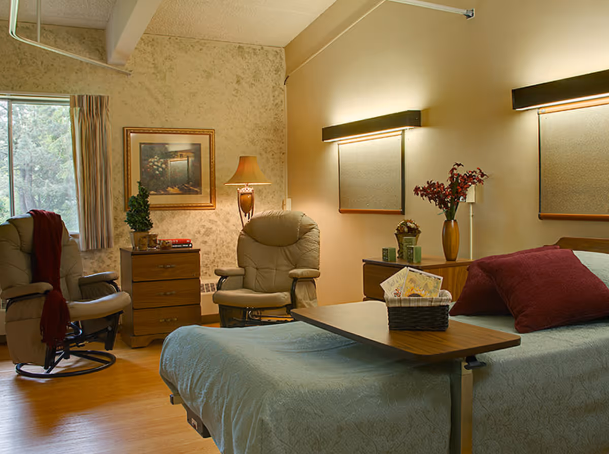 A cozy bedroom in a senior living facility with a bed covered in a light blue quilt and red pillows. There is a wooden overbed table with a basket containing cards and a small plant. Two beige recliner chairs are positioned near a window with curtains. A wooden nightstand with a lamp, a framed picture on the wall, and a vase with flowers add to the warm ambiance.
