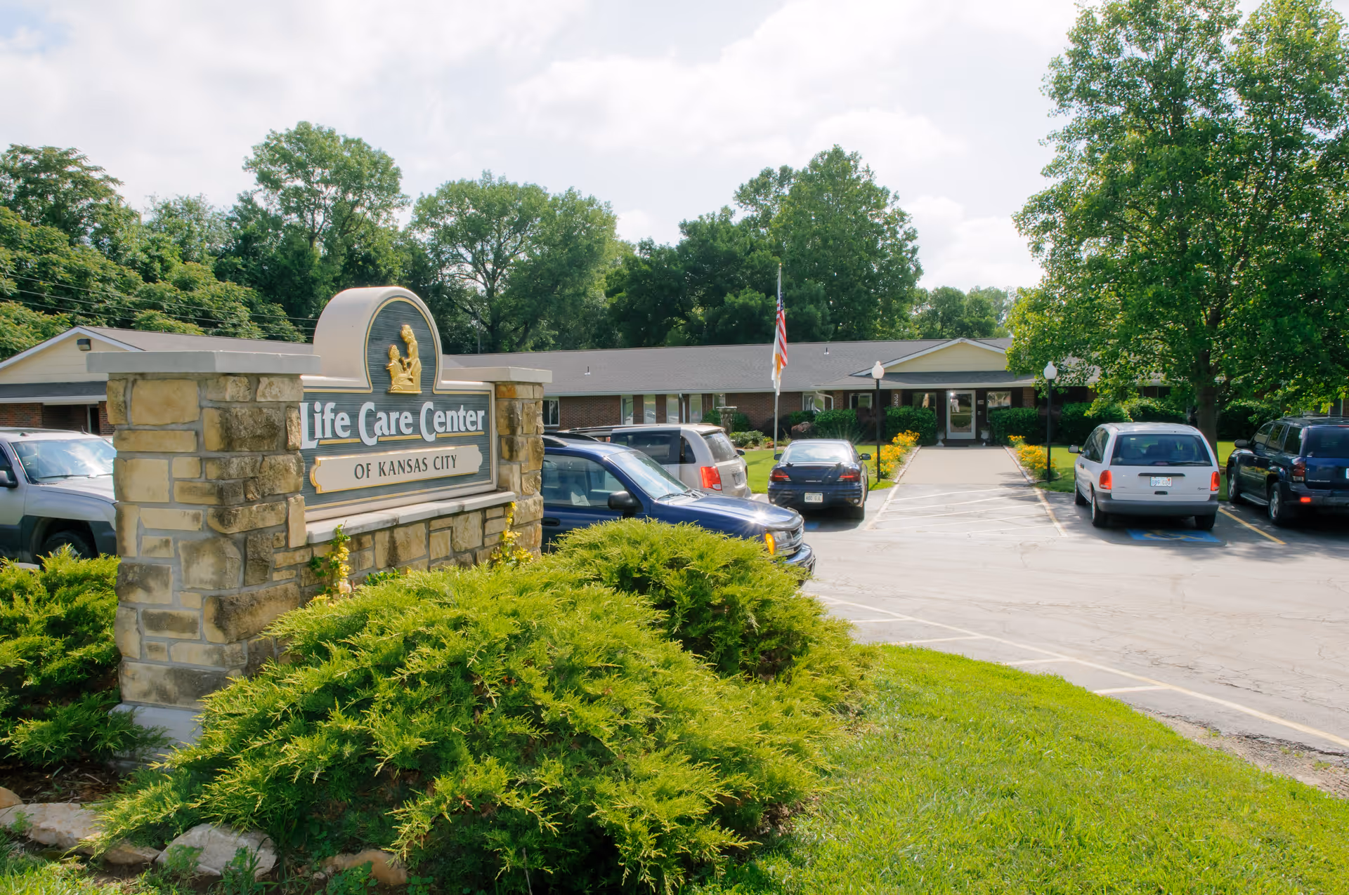 Stone sign reading "Life Care Center of Kansas City" in front of the facility building, parking lot, and landscaped shrubs.
