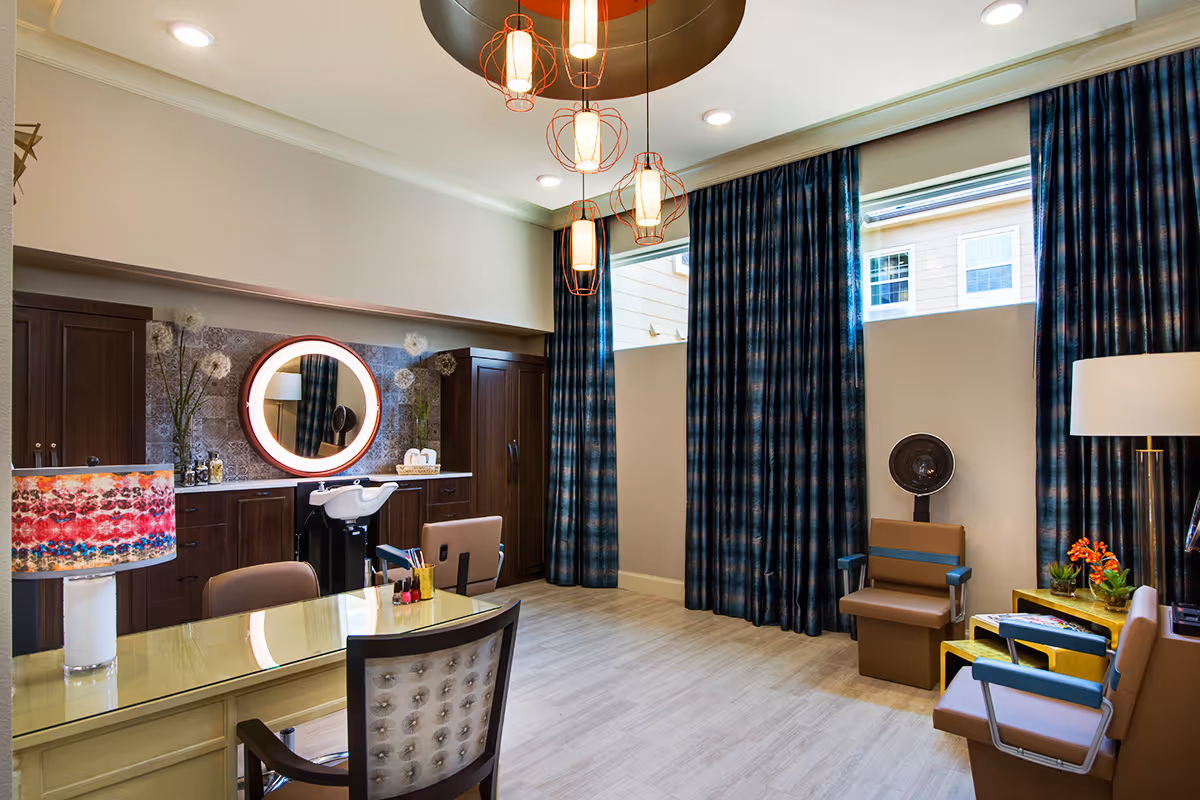 A bright and modern salon room with a large mirror surrounded by a circular light, dark wood cabinets, a salon chair, and a hair washing station. The room features a glass table with a chair, colorful lamp, and several brown chairs with blue armrests. Large windows with blue plaid curtains let in natural light, and there are decorative plants and flowers on the cabinets and tables.