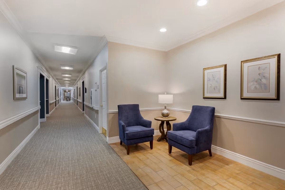 A long, well-lit hallway in a senior living facility with beige walls and carpeted floor. On the right side near the foreground, there are two blue upholstered armchairs with a small round wooden table between them, topped with a lamp. Two framed botanical prints hang on the wall above the chairs.