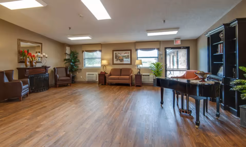 A spacious and well-lit common area with wooden flooring, featuring a black grand piano on the right side, a dark wooden bookshelf, a brown loveseat and two armchairs, a decorative fireplace with a mirror above it, potted plants, and two windows letting in natural light.