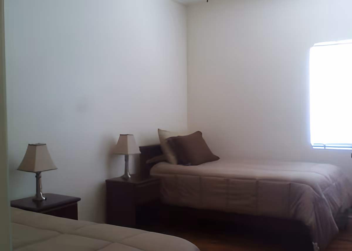 A simple bedroom with two single beds, each with beige bedding and a brown pillow. There are two bedside tables, each with a lamp. A window with bright light is on the right side of the room, and the walls are plain white.