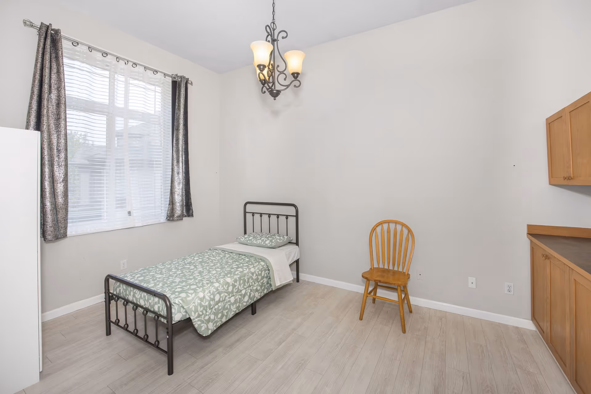 Simple bedroom with a single metal bed, wooden chair, window with curtains, and built-in wooden cabinets.