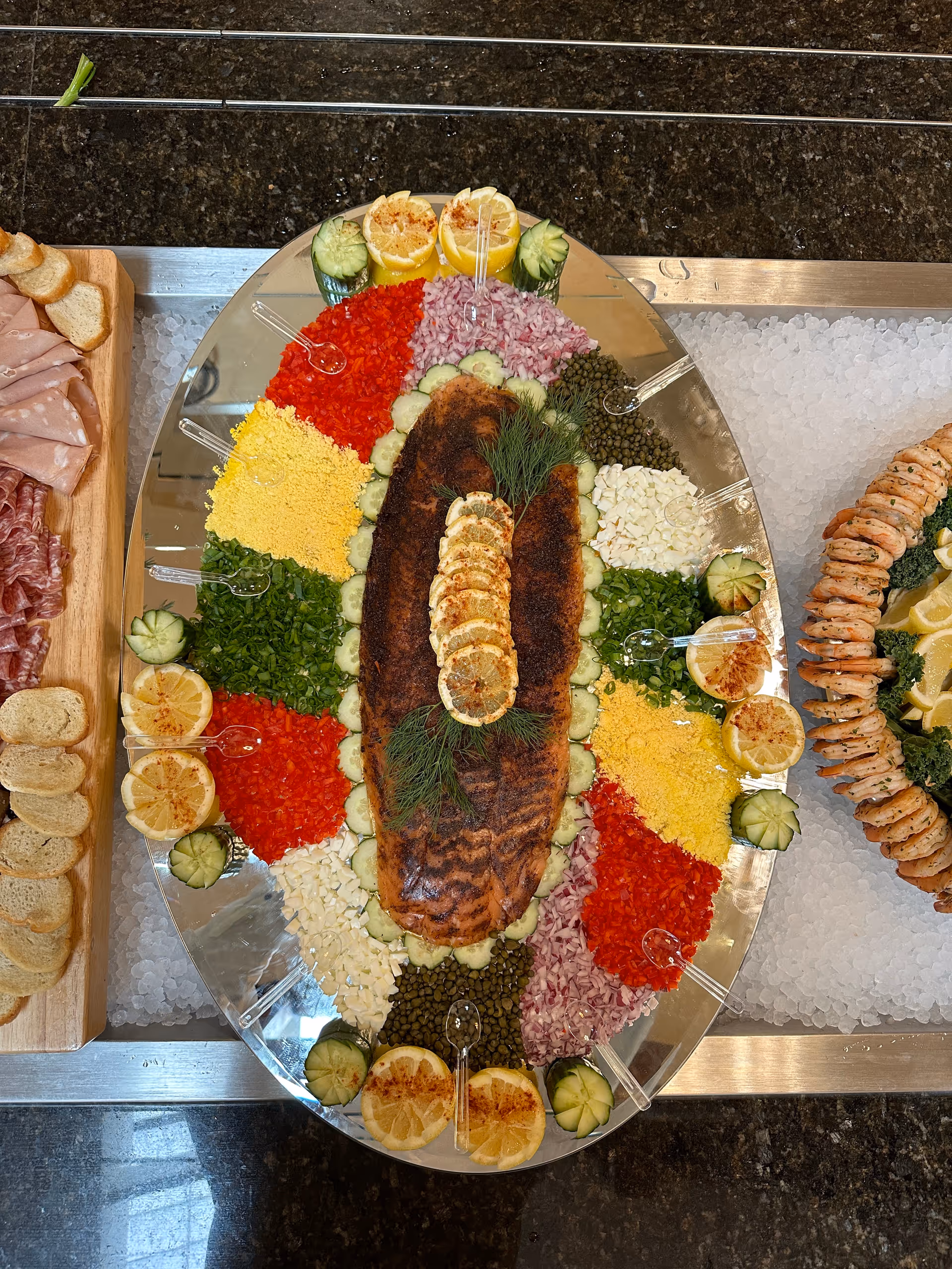 A large oval platter with a whole cooked fish garnished with lemon slices and dill in the center, surrounded by neatly arranged sections of chopped vegetables and garnishes including red peppers, chopped onions, capers, chopped eggs, and cucumber slices. The platter is placed on a bed of ice on a dark granite countertop, with additional platters of sliced bread and shrimp cocktail visible on either side.
