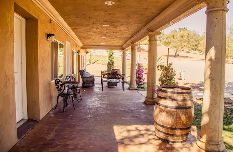 Covered outdoor patio area with stone columns, a wooden barrel, metal chairs and tables, a wicker chair with cushions, and flowering plants along the edge. The patio overlooks a dry, grassy landscape with trees and a road in the background.