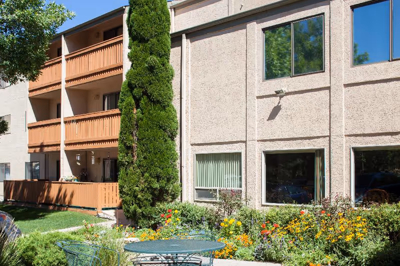 Outdoor garden area at Mountain View Independent Living with colorful flowers, green shrubs, a tall tree, and a metal table with chairs. The background shows a beige building with balconies and windows reflecting the blue sky.